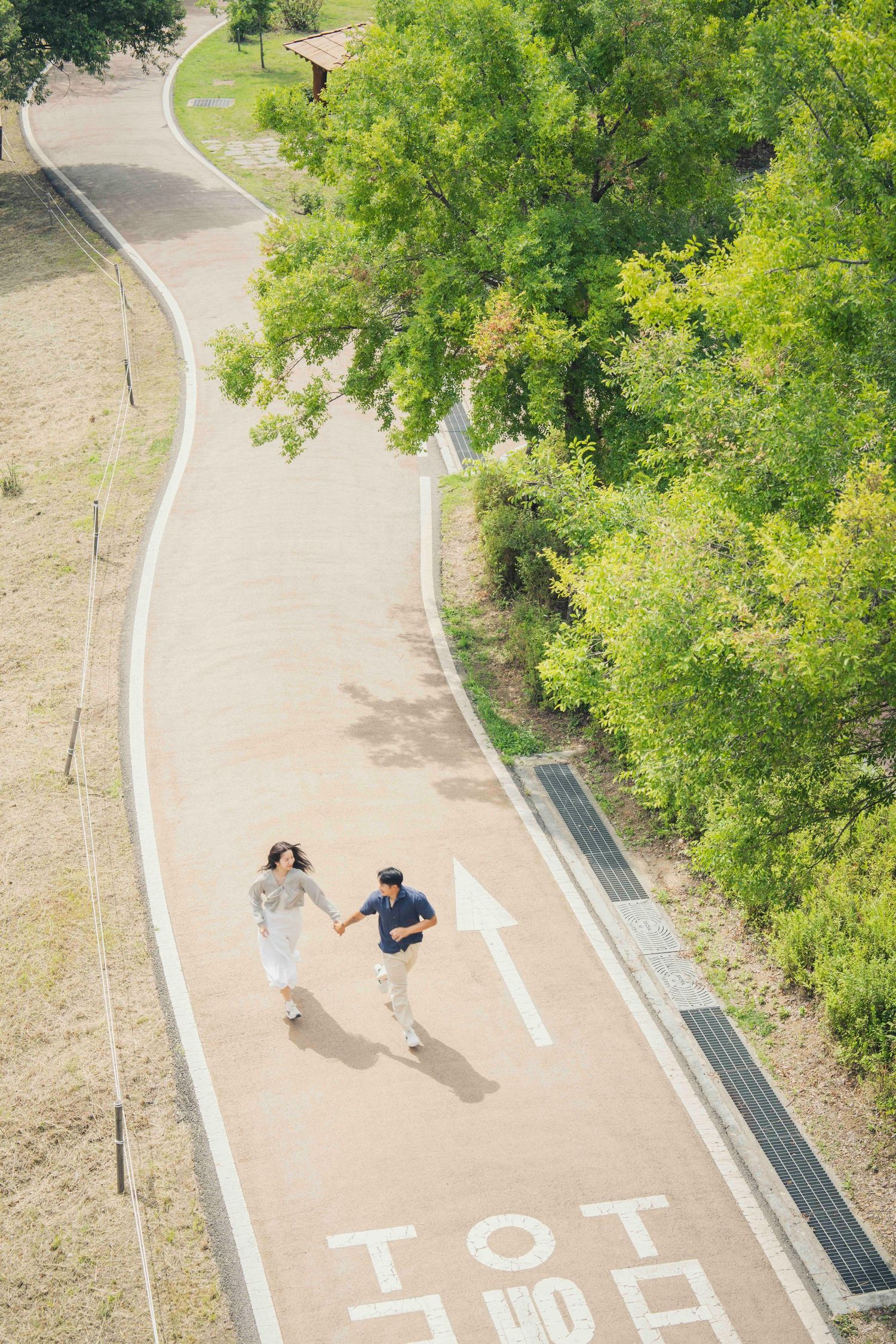 Aerial view of a couple holding hands and running along a curved park pathway surrounded by lush green trees.