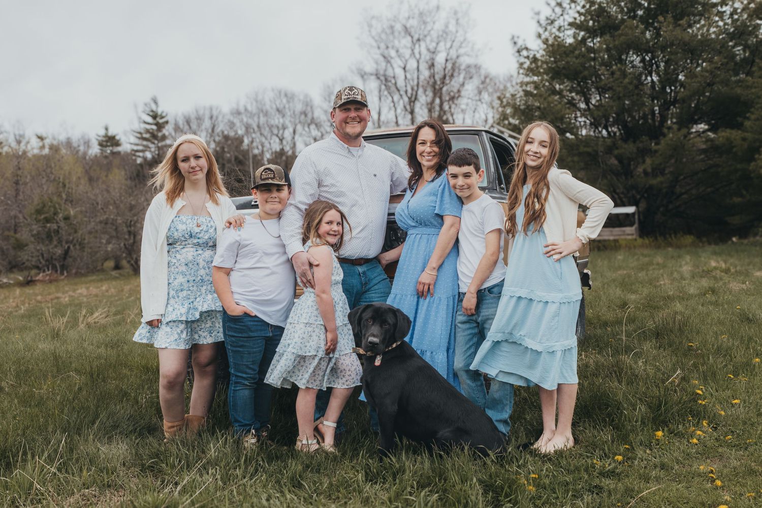 Family of six poses with black lab dog in grassy field near truck, wearing coordinated blue and white outfits.
