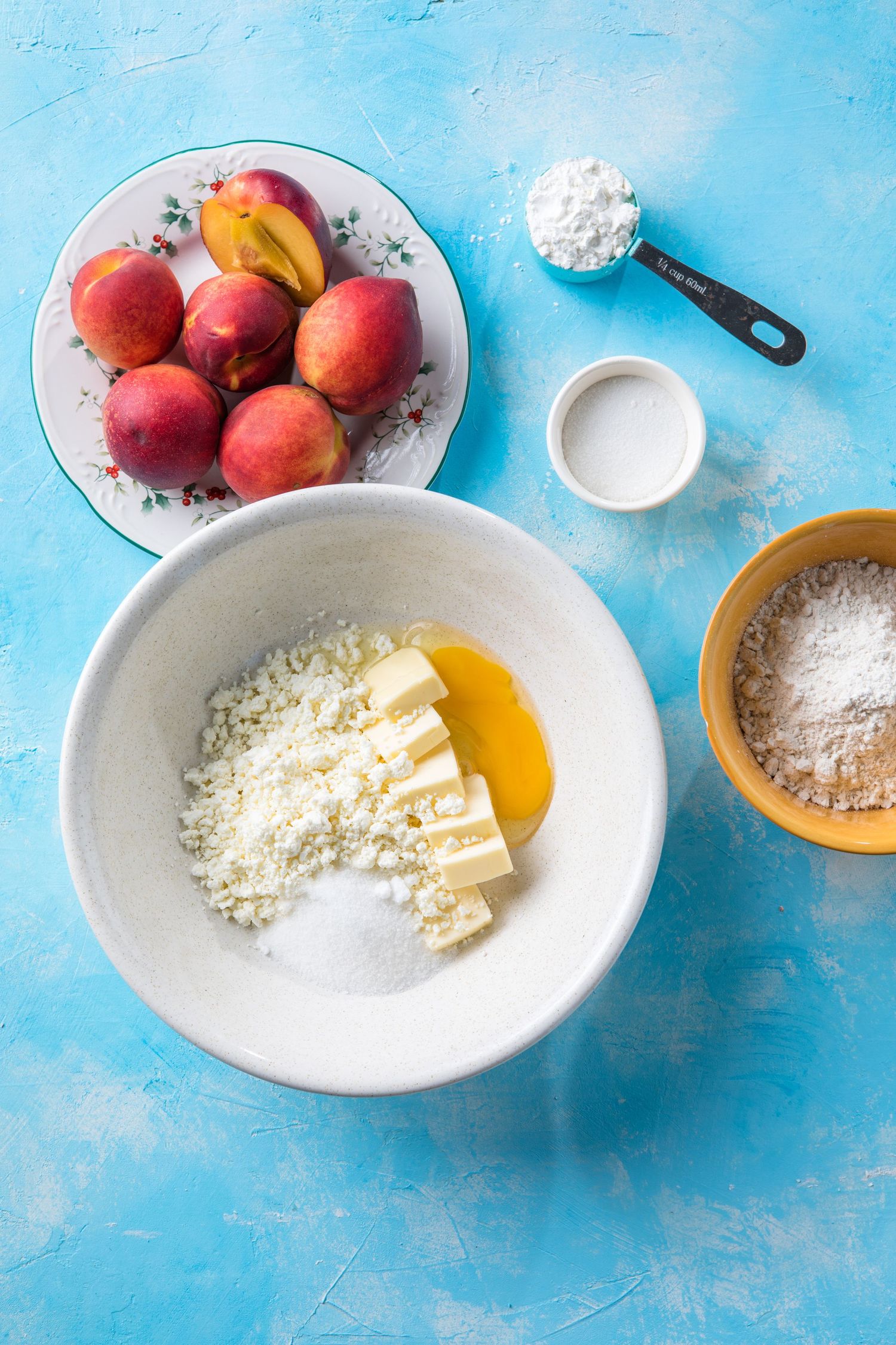 Fresh peaches, butter, eggs, flour, and sugar ingredients laid out on a blue surface for a peach dessert recipe.