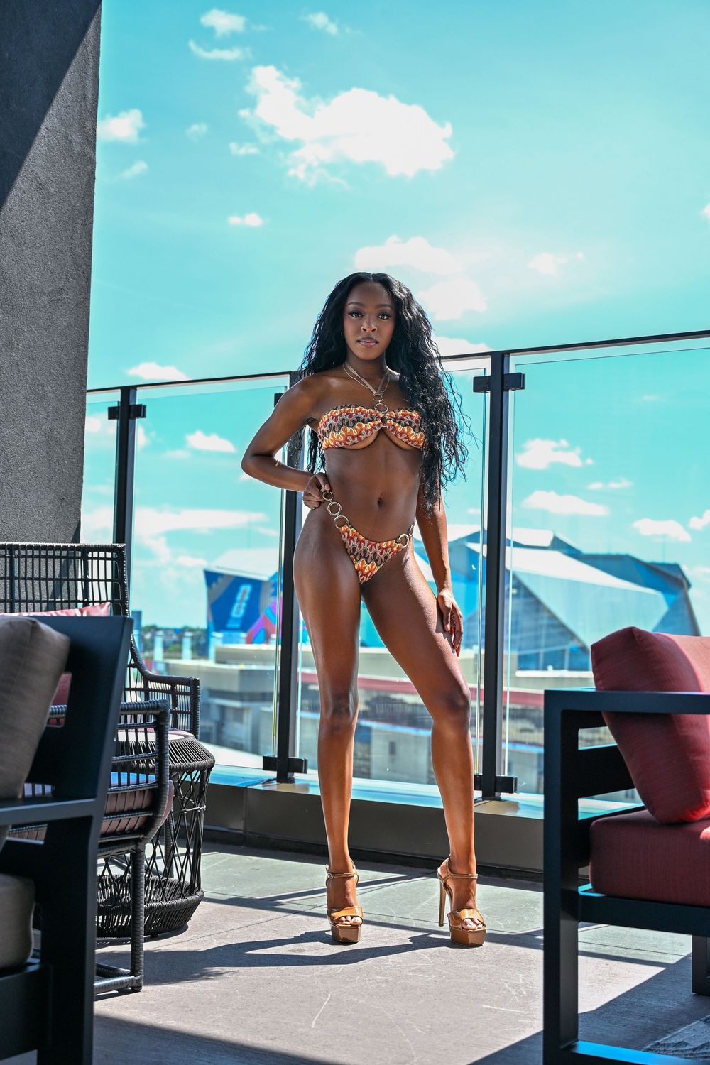 A confident woman in a leopard print bikini and heels poses on a rooftop terrace under a bright blue sky.
