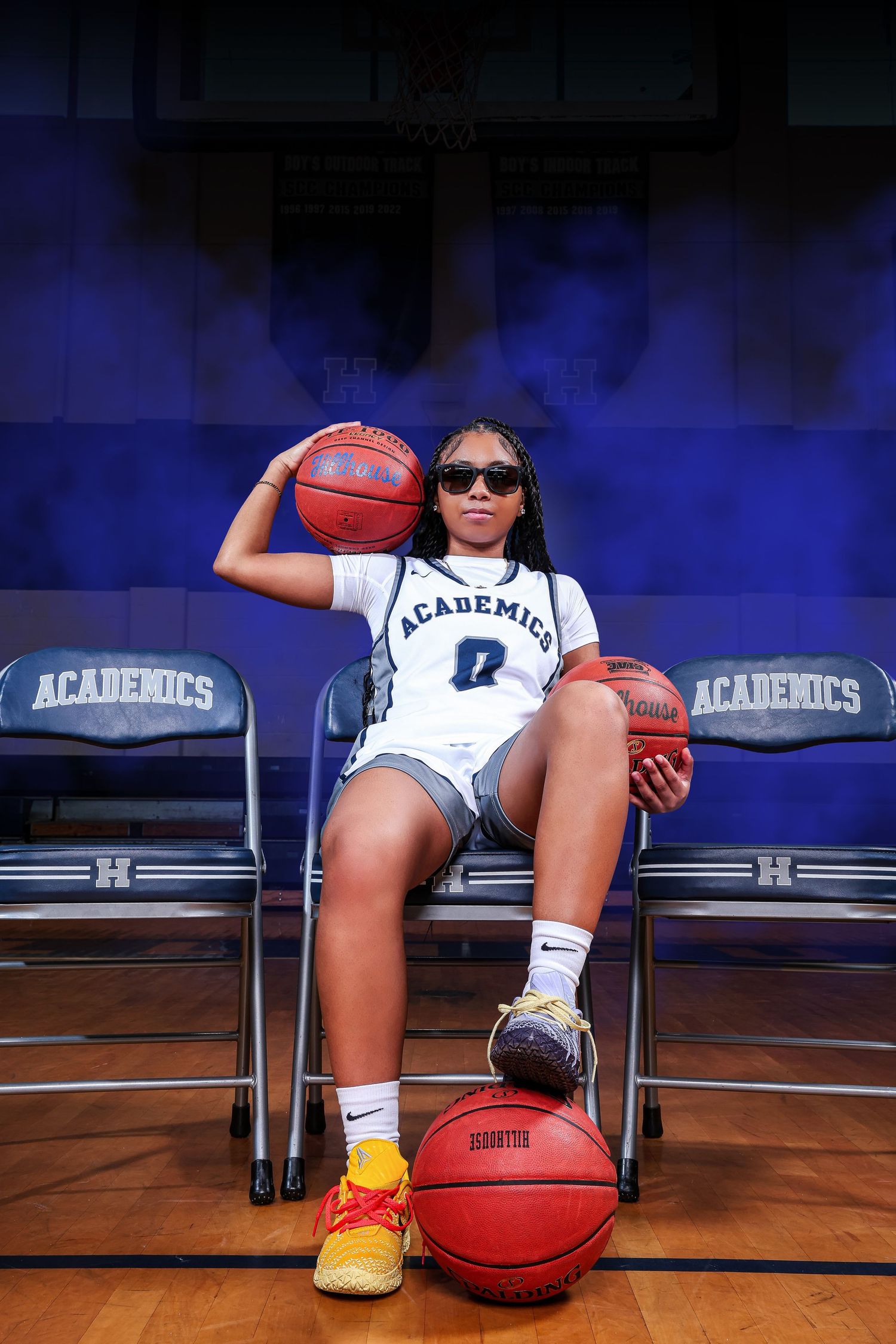 Female basketball player in Academicos jersey #0, posing confidently with multiple basketballs on gym bleachers wearing sunglasses.