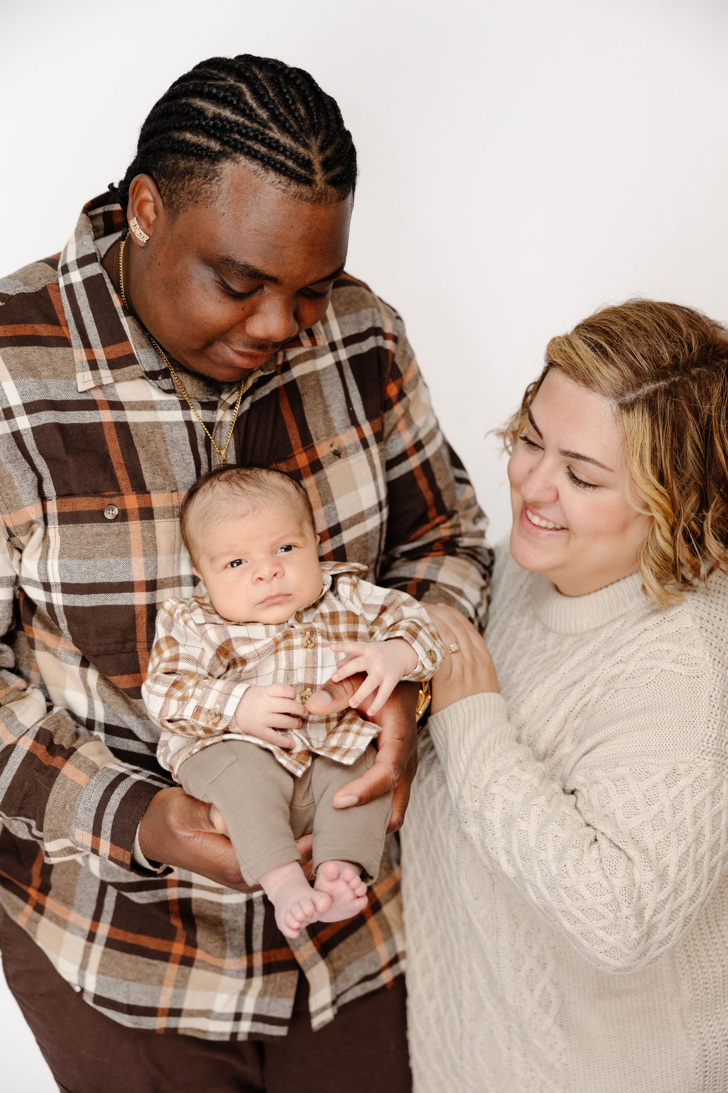 A joyful interracial couple holds their newborn baby, dressed in matching plaid outfits, against a white background.