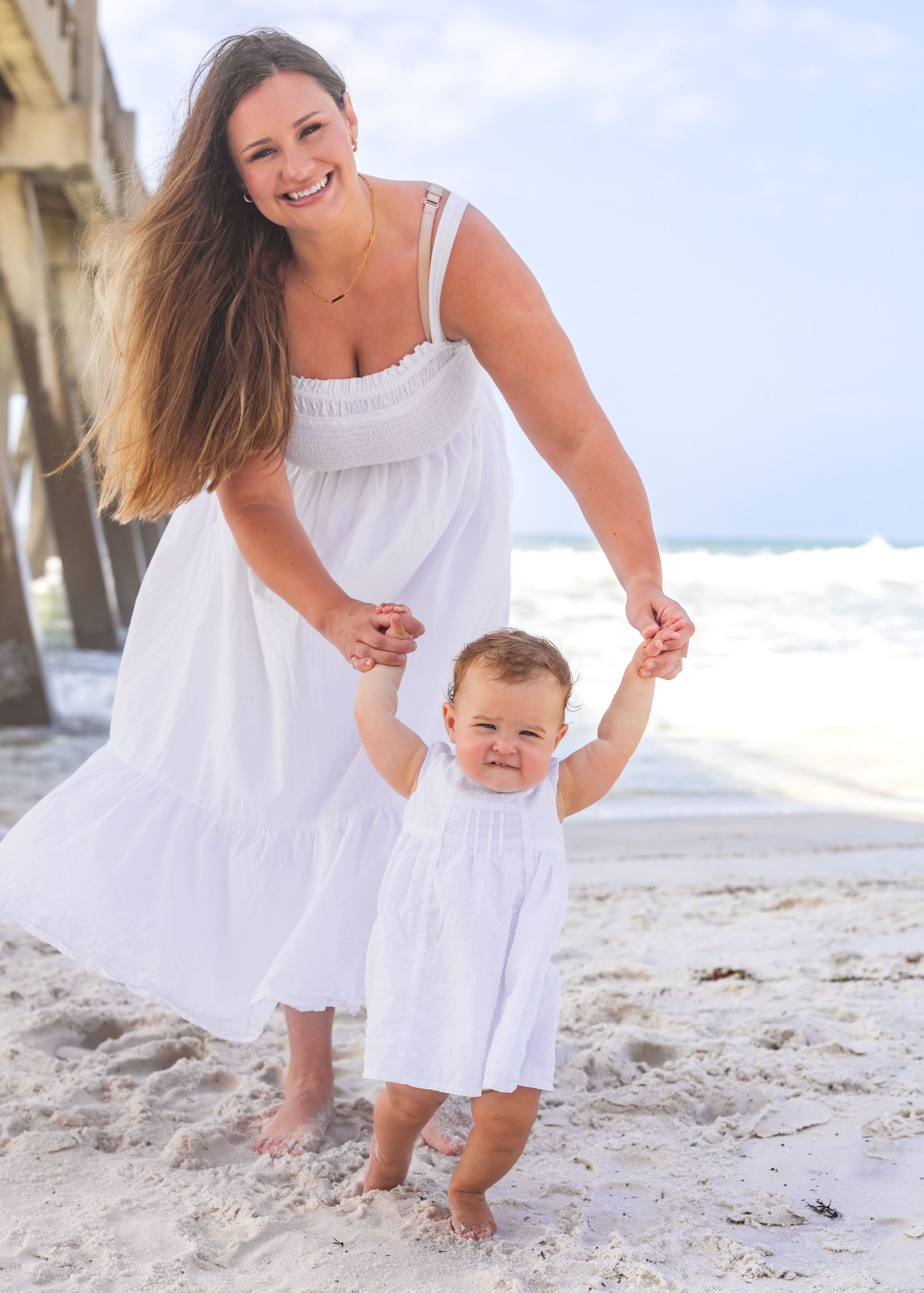A smiling mother in a white dress helps her baby girl walk on a sunny beach near a pier.