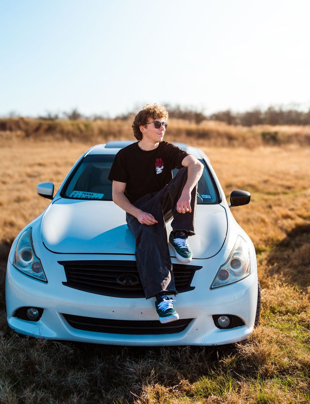 Young man in sunglasses sitting on white Infiniti sports car hood in sunny field wearing black outfit and blue sneakers.