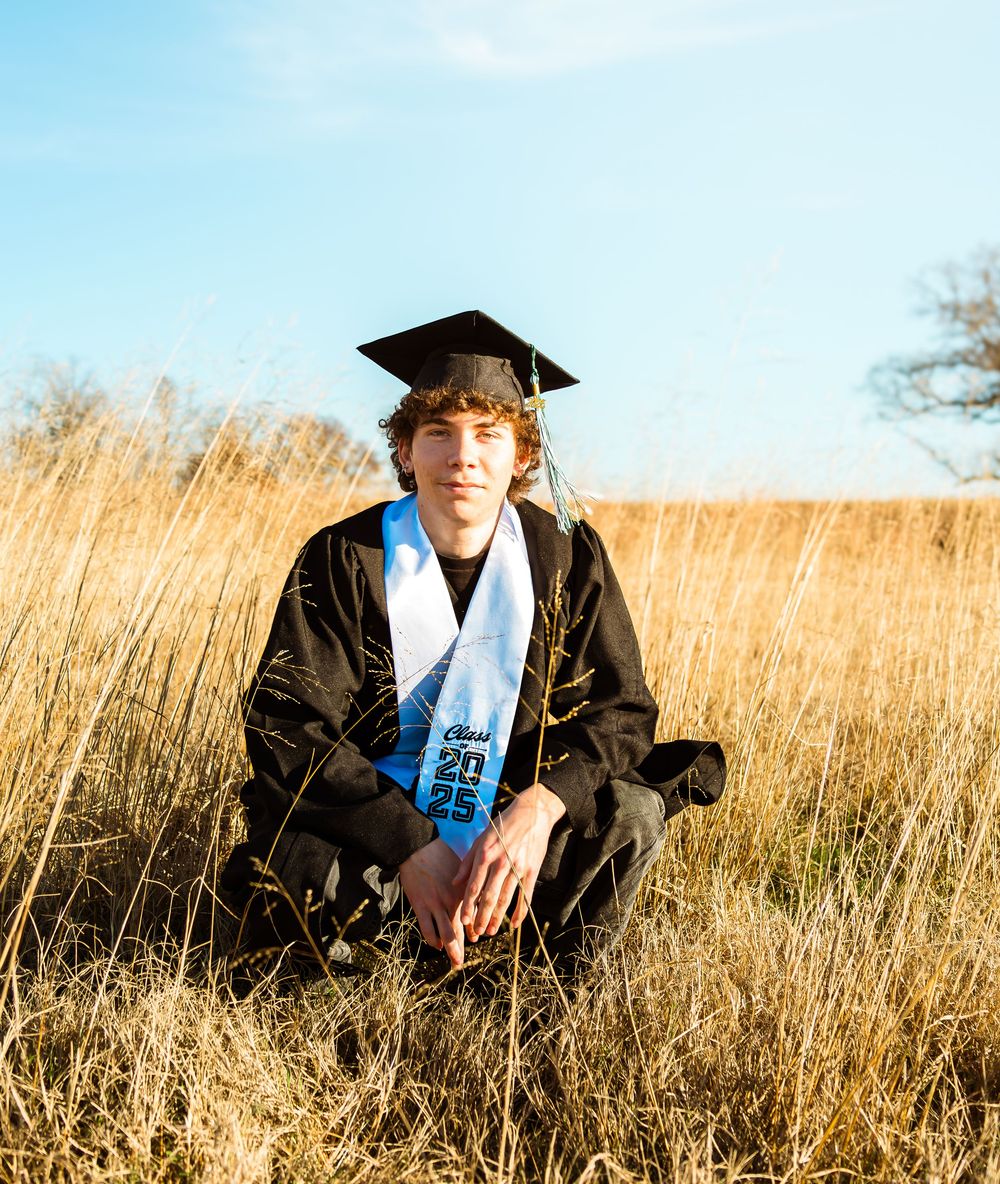 A young man in graduation cap and gown sits in a golden field wearing a blue sash.