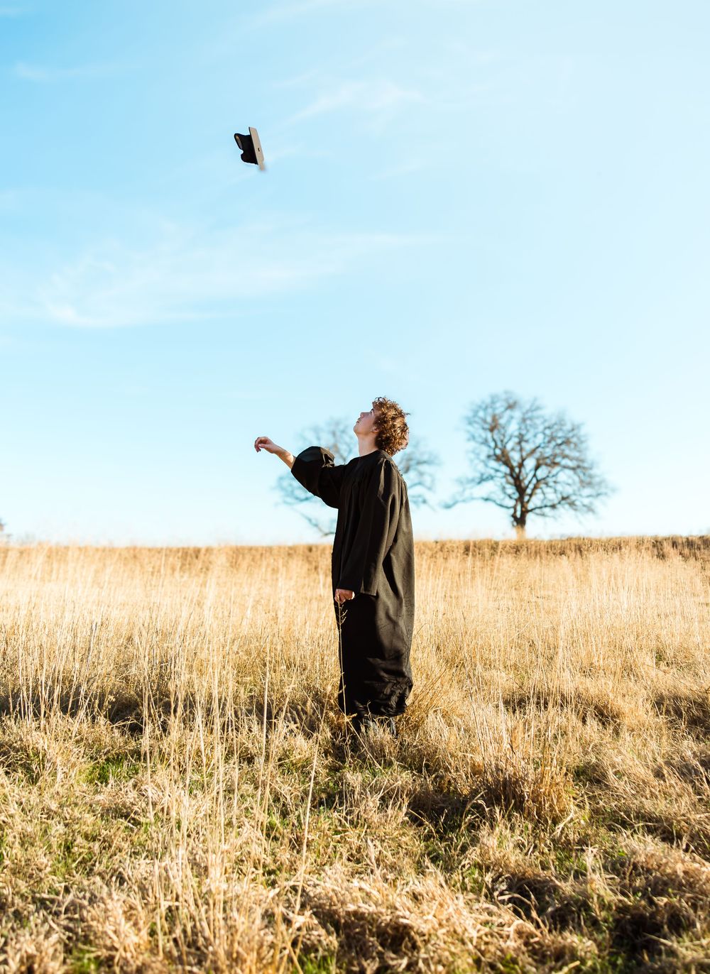A graduate in black gown tosses cap into blue sky above a golden meadow.