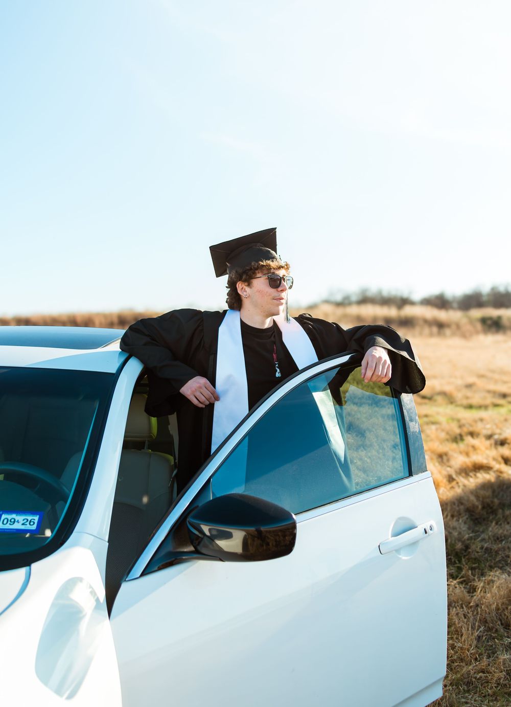 Graduate in cap and gown leaning on white car door in open field on a clear sunny day.