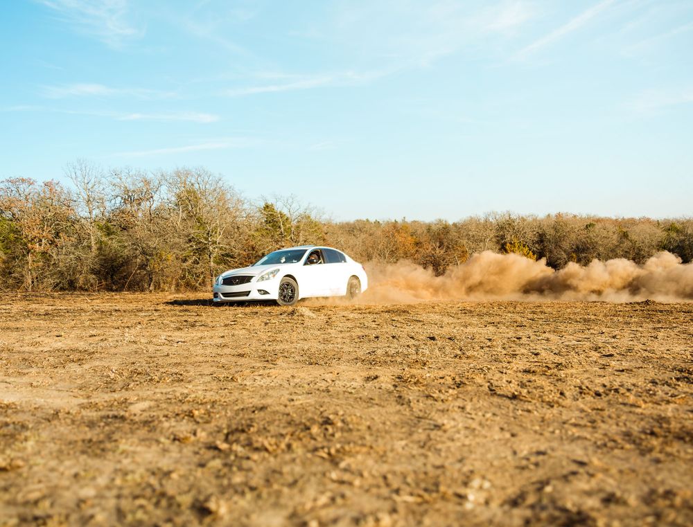 White sedan drifting on dirt field, kicking up dust clouds, surrounded by autumn trees under blue sky.