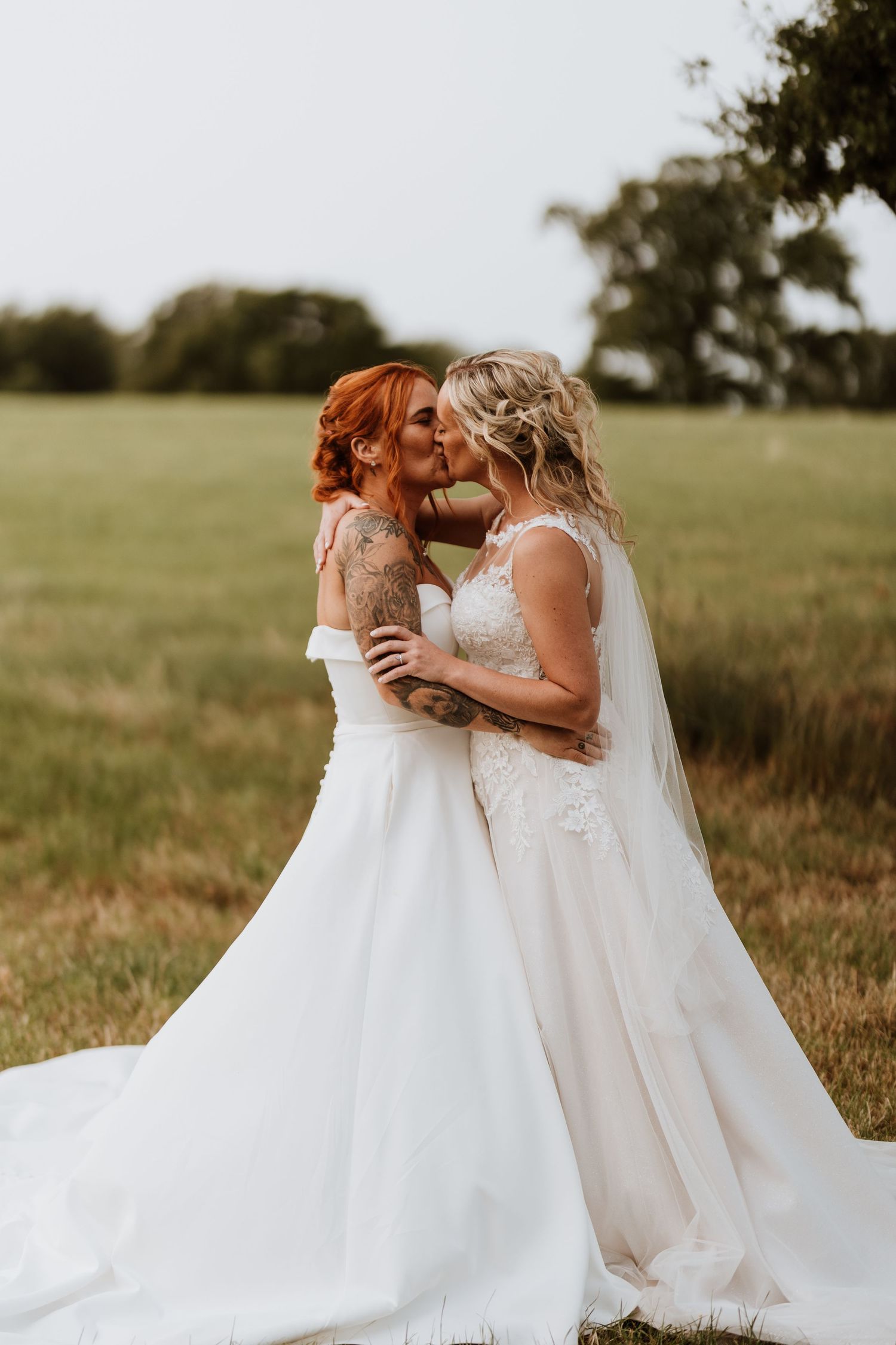 Two brides in white wedding gowns share a romantic kiss in a lush green field on their wedding day.
