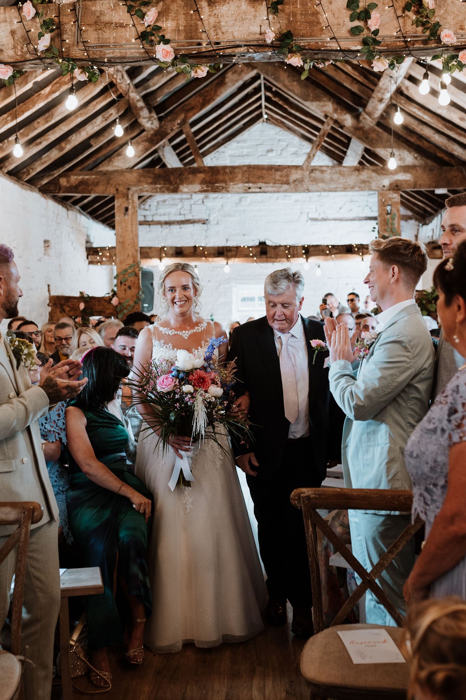 Bride walks down the aisle in a rustic barn wedding venue decorated with fairy lights and pink floral garlands.