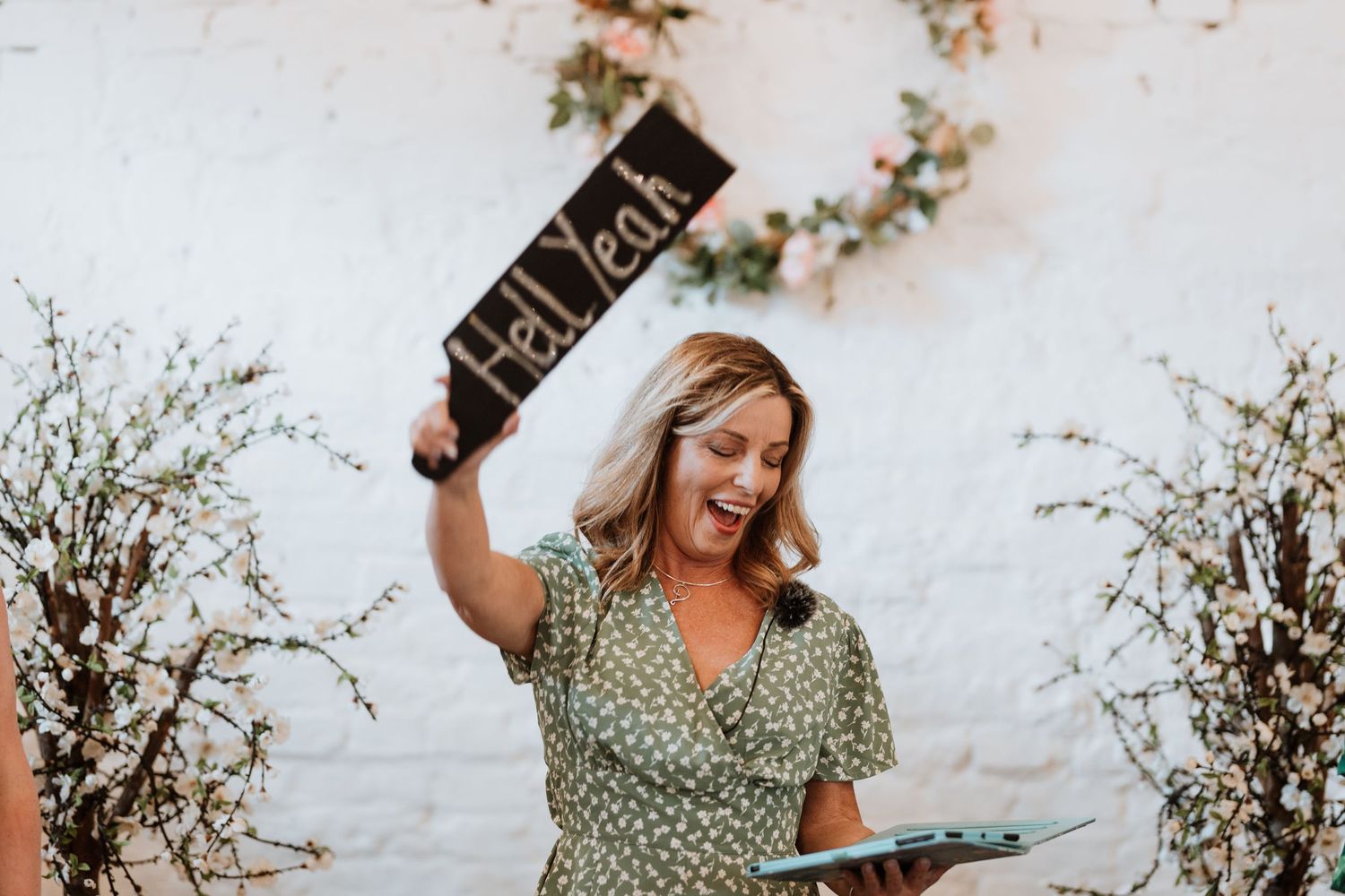Woman in green floral dress holding a Hell Yeah sign at a wedding celebration with floral arch backdrop.