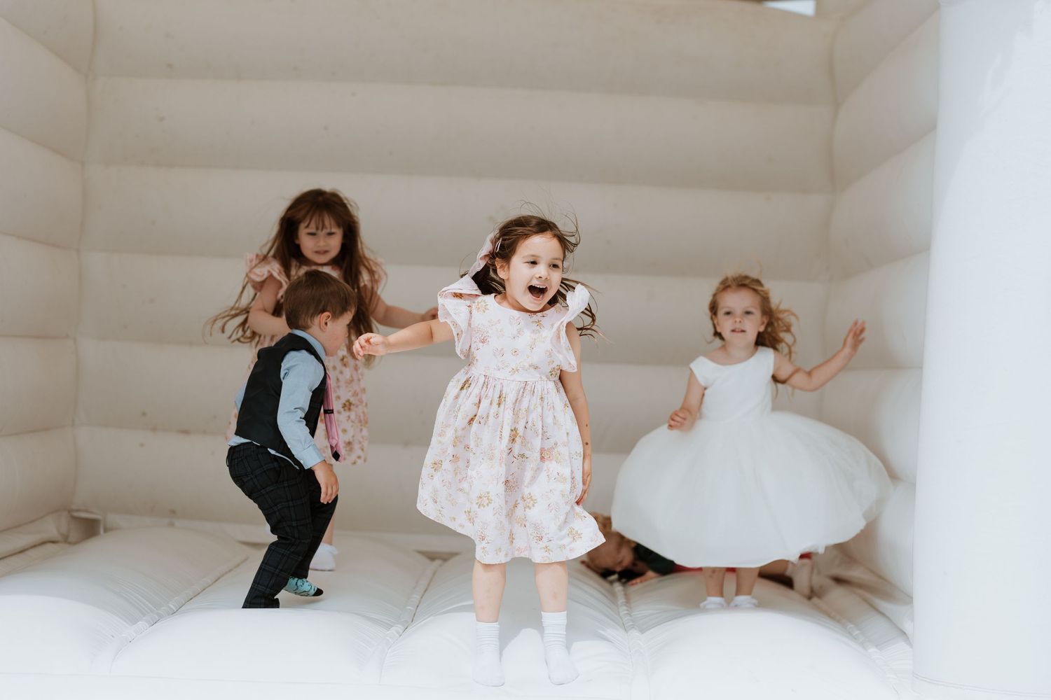 Four children playing and jumping joyfully inside a white bouncy castle at a wedding reception.