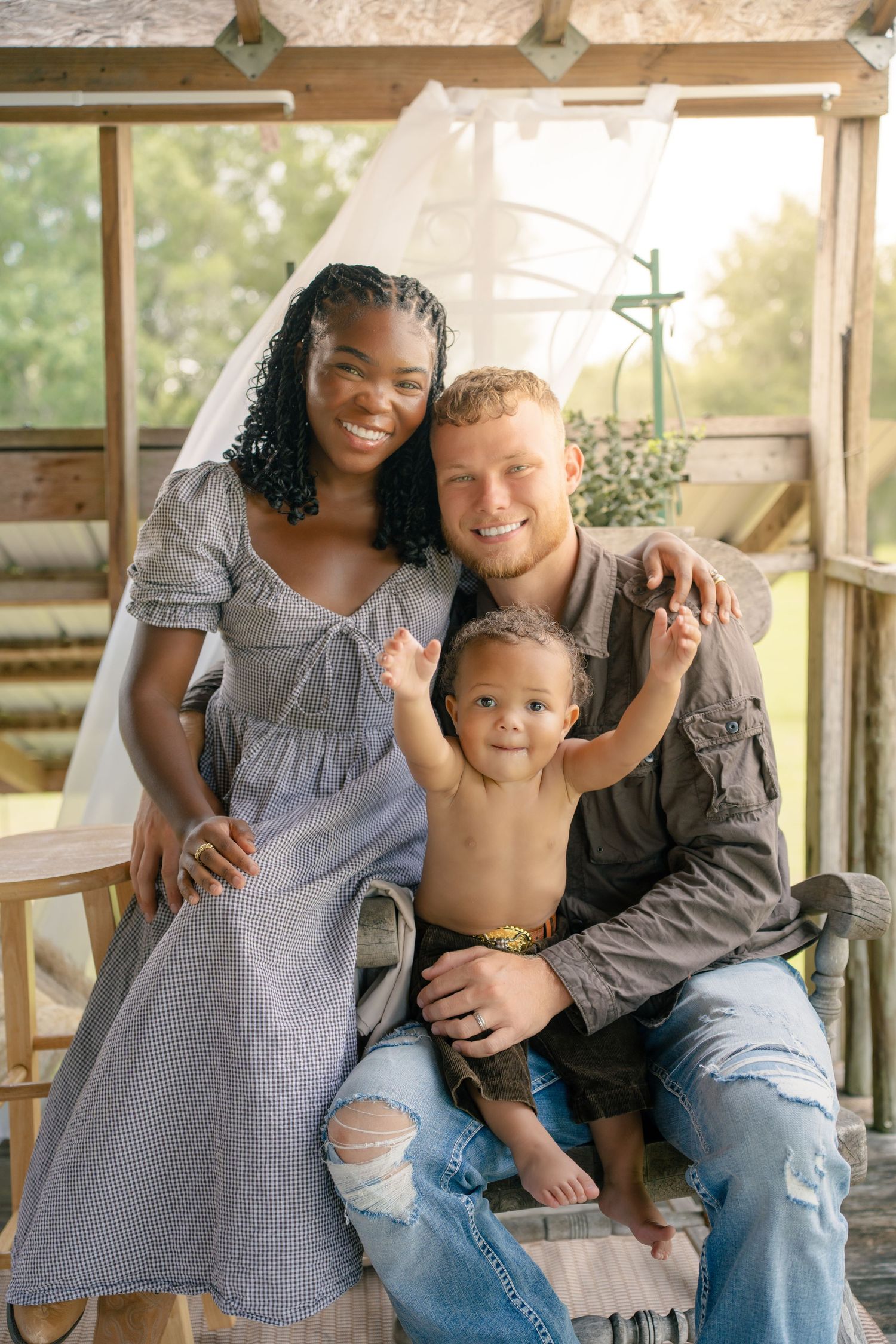 Interracial family portrait with smiling mother, father, and baby on a rustic porch surrounded by greenery.