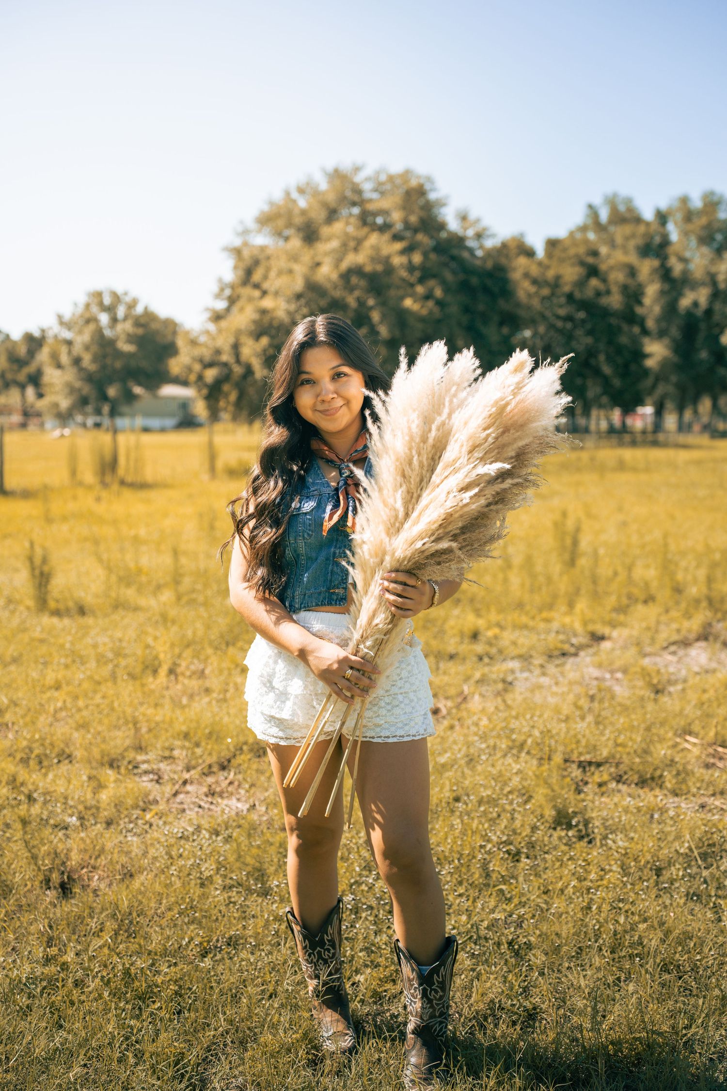 Young woman in denim vest, white shorts, and cowboy boots holding pampas grass in a sunny green field.