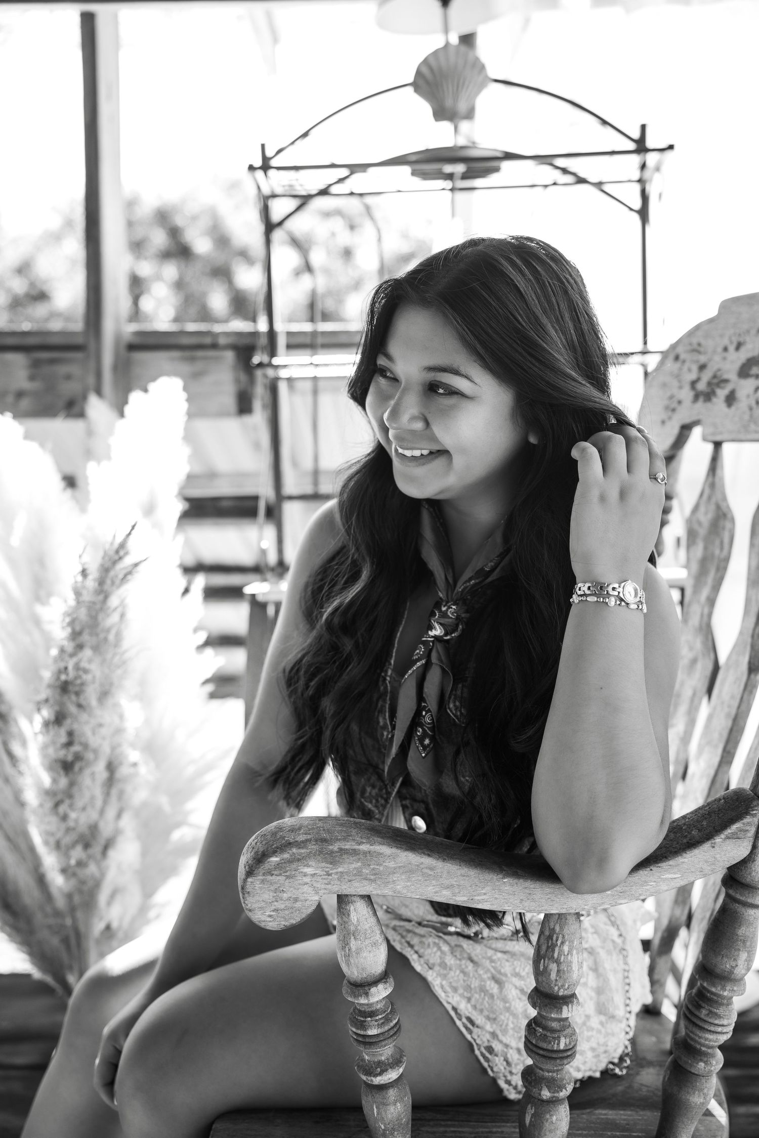 Black and white portrait of a smiling young woman with long wavy hair sitting in a wooden chair.