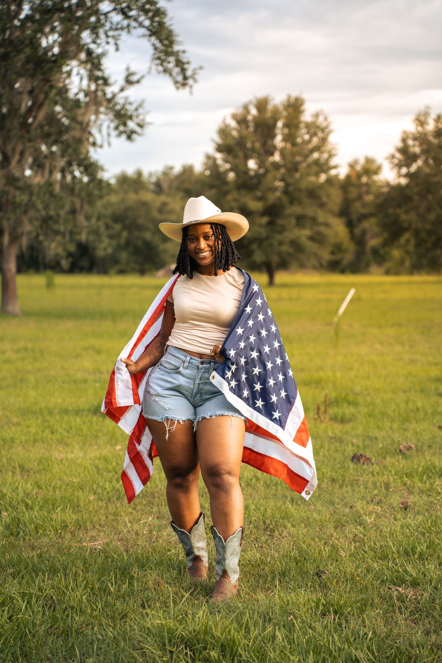 Young Black woman in cowboy hat and boots holds American flag in sunny green field.