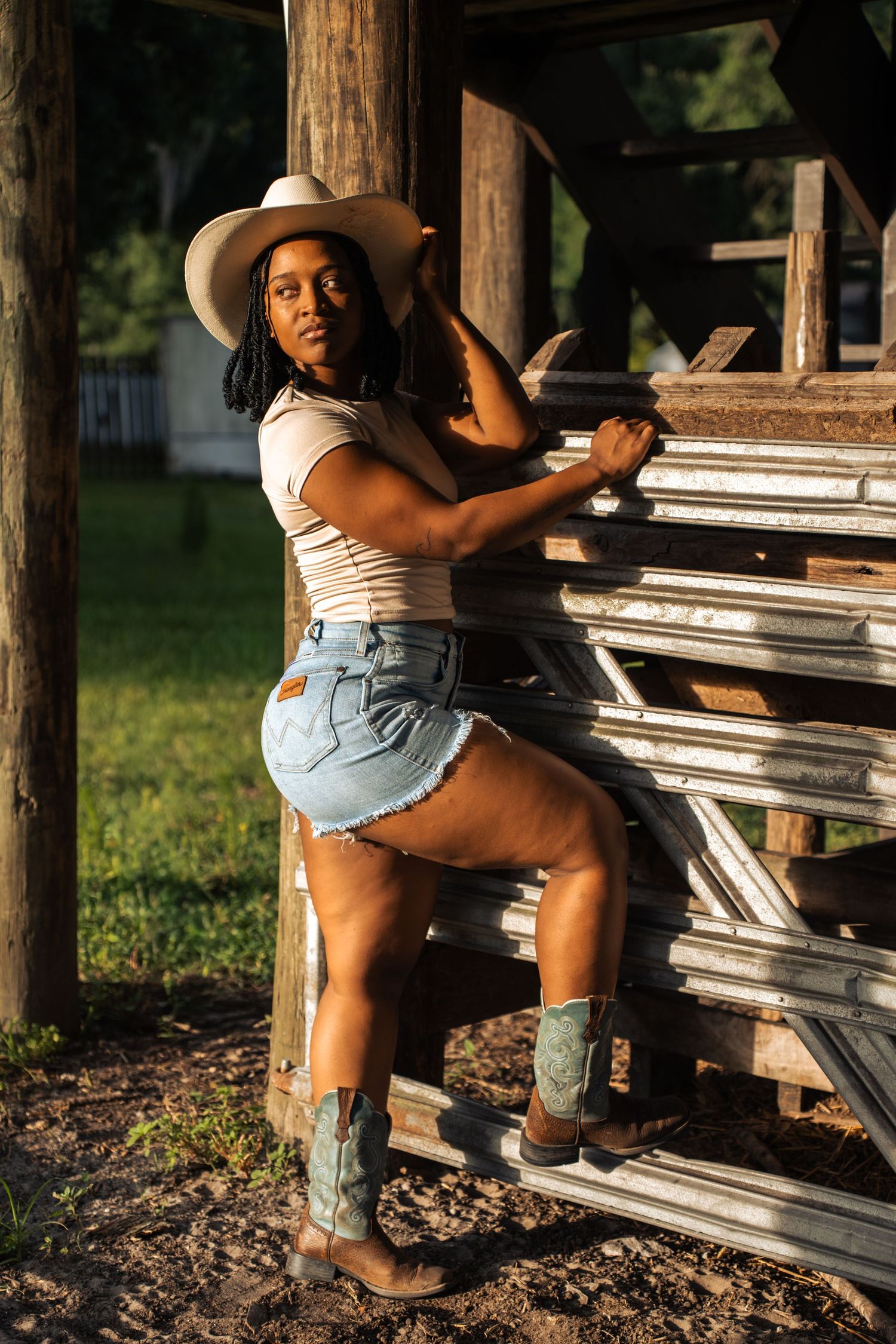 A Black woman in cowboy hat, denim shorts, and boots poses against rustic wooden structure in golden hour light.