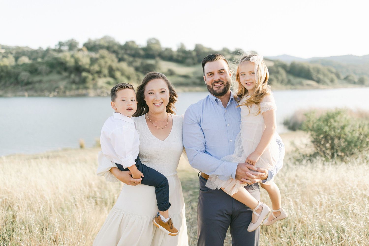 Happy family of four posing outdoors near a lake, parents holding two young children in a sunny field.
