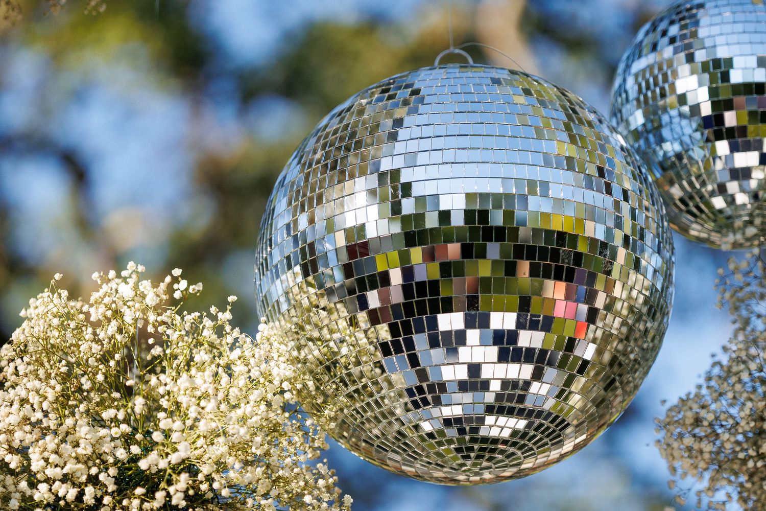 Shiny disco mirror balls hanging outdoors among white flowers with a blurred blue sky background.