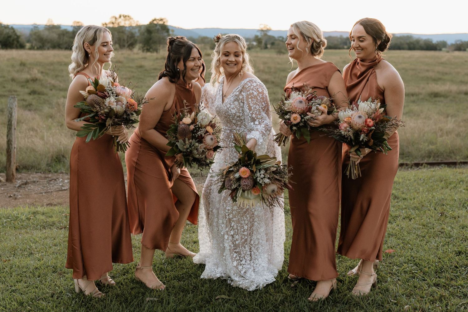 Bride in lace gown with four bridesmaids in rust satin dresses holding bouquets in a green field.