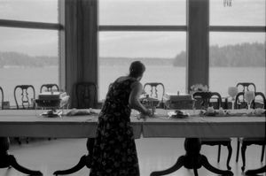 Woman setting a long dining table by large windows overlooking a serene lake, black and white photography.