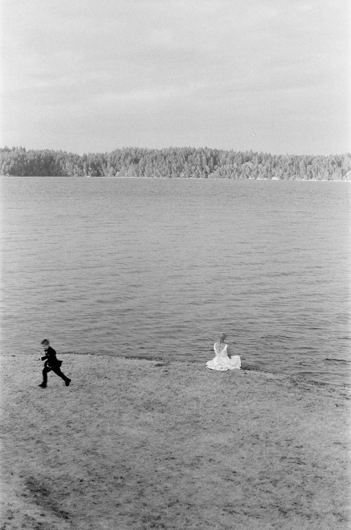 Black and white photo of a child running toward a person in white dress sitting by a vast lake.