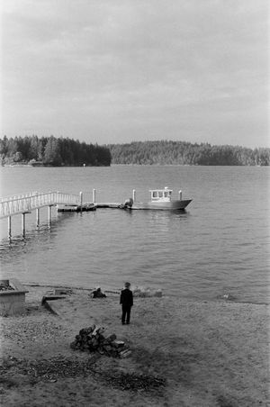 Black and white photo of a man watching a boat dock at a quiet lakeside pier.