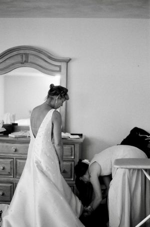 Black and white photo of a bride in a backless wedding gown standing near a dresser while a bridesmaid adjusts her dress.