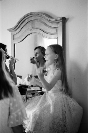 Young girl in white dress applies makeup at vanity mirror in black and white photo.