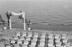 Black and white photo of empty wedding chairs facing a floral arch by the water's edge.