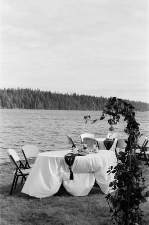 Black and white photo of an elegant outdoor lakeside wedding reception table with white linens and floral arch.