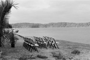 Rows of folding chairs arranged outdoors by a calm lake with forested shoreline in black and white.