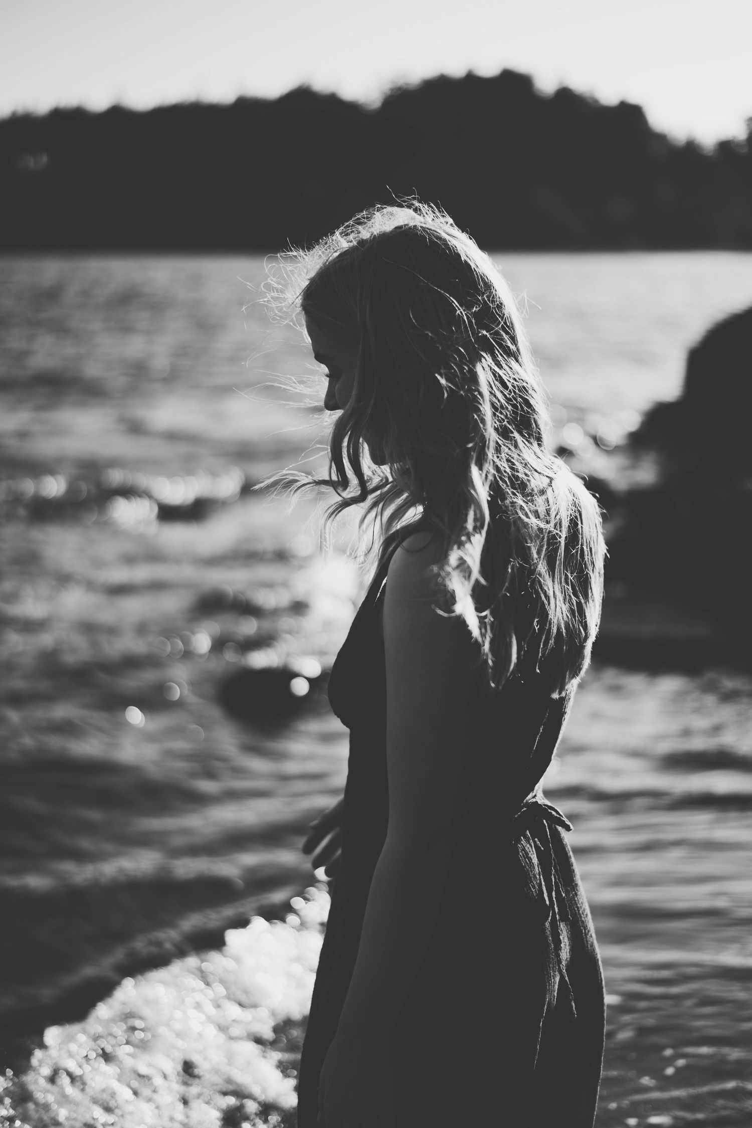 Black and white photo of a woman with long hair standing by the ocean shore in a flowing dress.