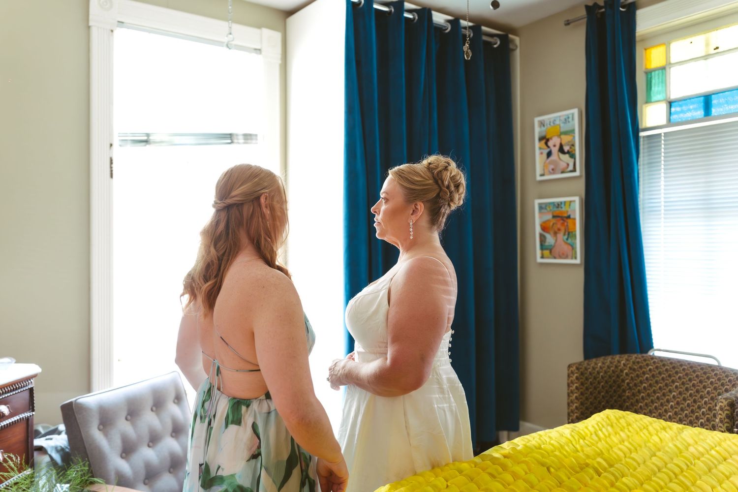 Bride in white gown holds hands with bridesmaid in floral dress inside a room with teal curtains.