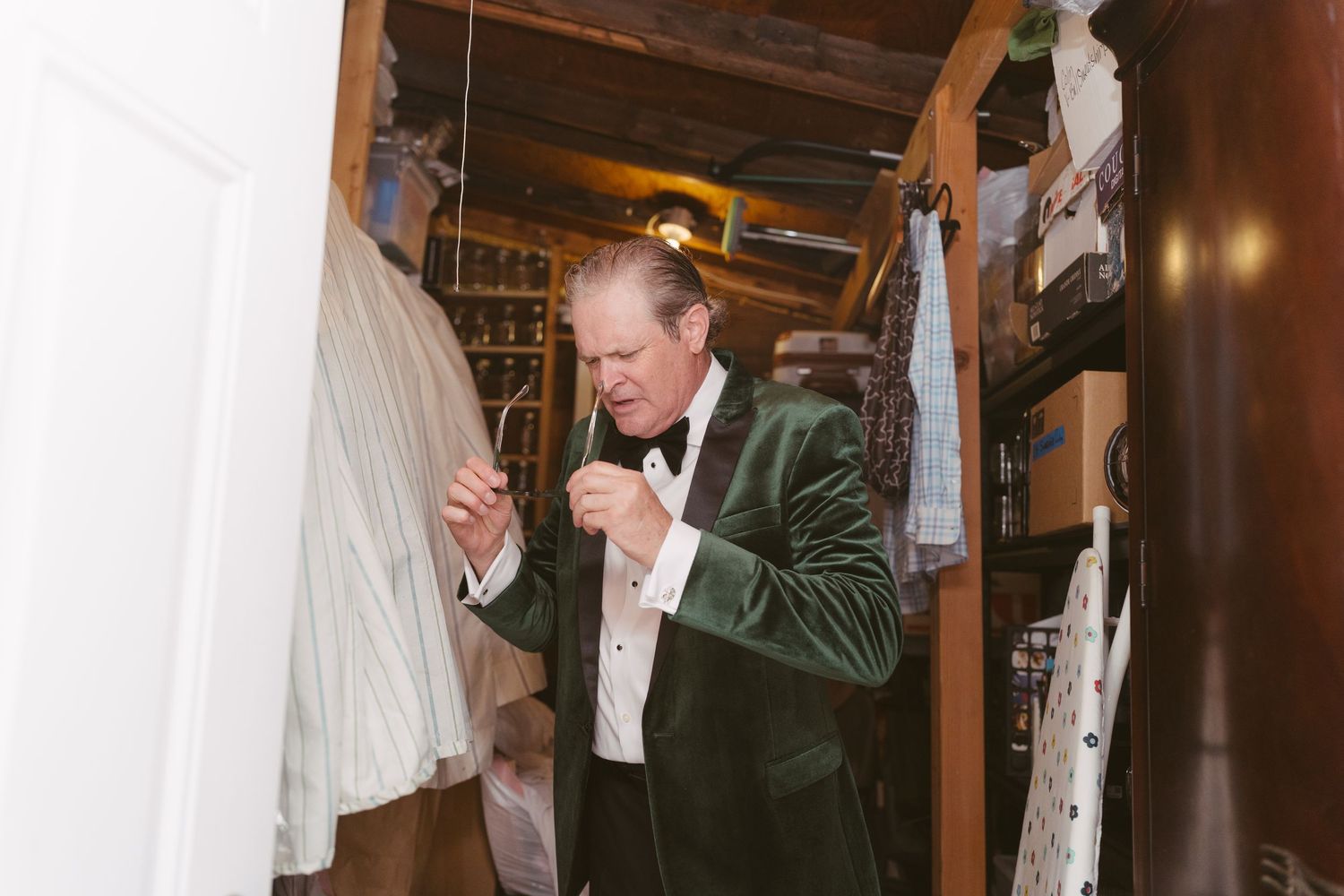 A man in a green velvet tuxedo jacket adjusts his bow tie in a closet before a wedding.
