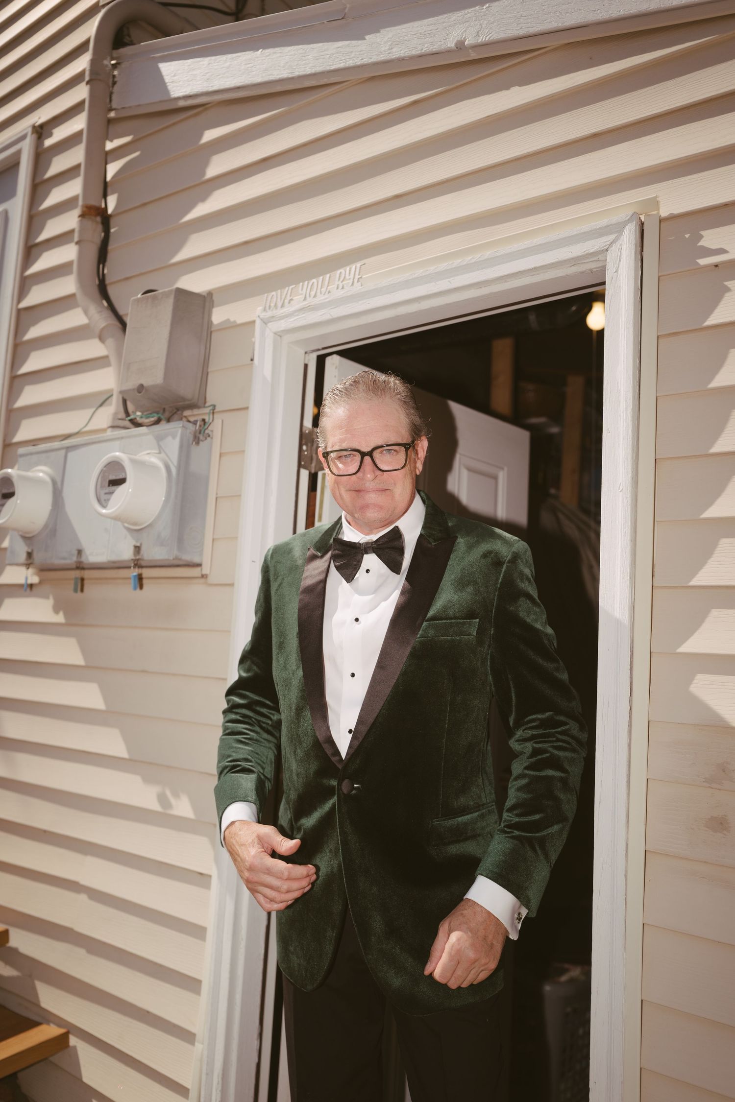 Elegant man in green velvet tuxedo with bow tie stands in doorway of white clapboard building.