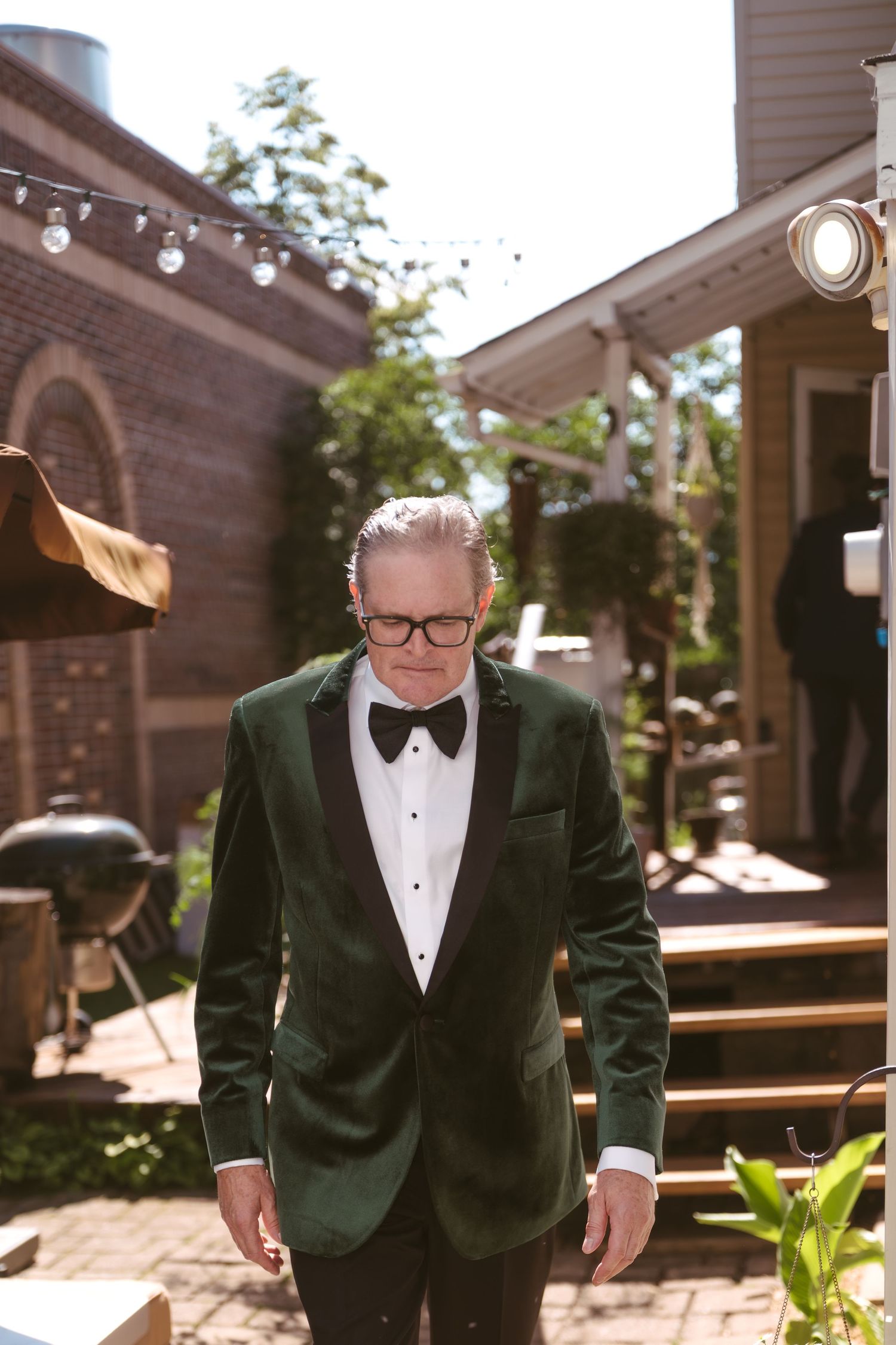 Distinguished man in green velvet tuxedo and bow tie walks through sunlit outdoor wedding venue.