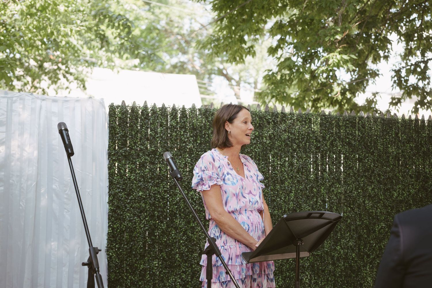 Woman in floral dress speaks at outdoor wedding ceremony podium in front of green hedge backdrop.