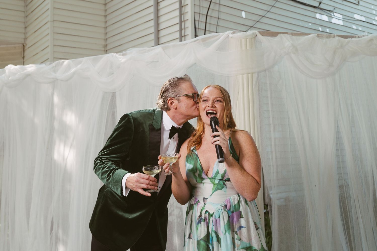 Groom in green velvet tuxedo kisses woman giving wedding toast holding champagne glass.