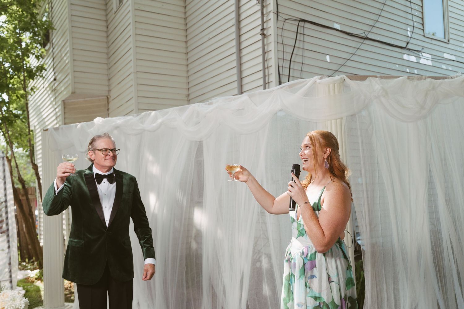Couple in formal attire raise champagne glasses during outdoor wedding reception toast.