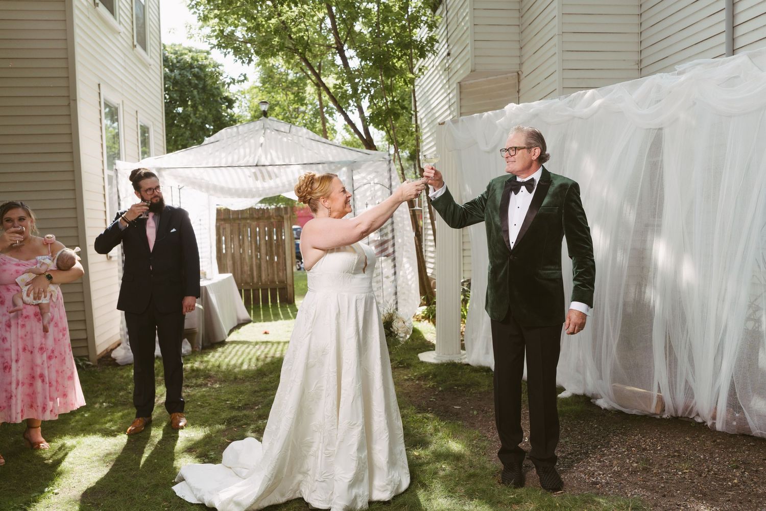 Bride and groom celebrate after ceremony, raising hands joyfully in backyard wedding venue with guests nearby.