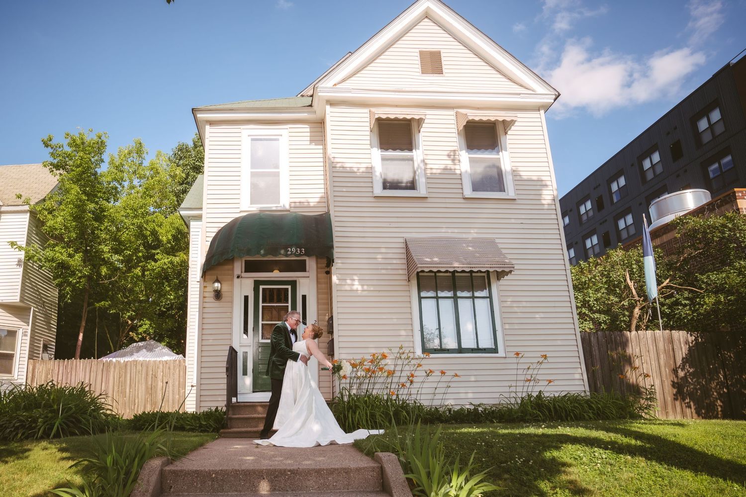 A bride and groom embrace in front of a charming white two-story house on a sunny day.