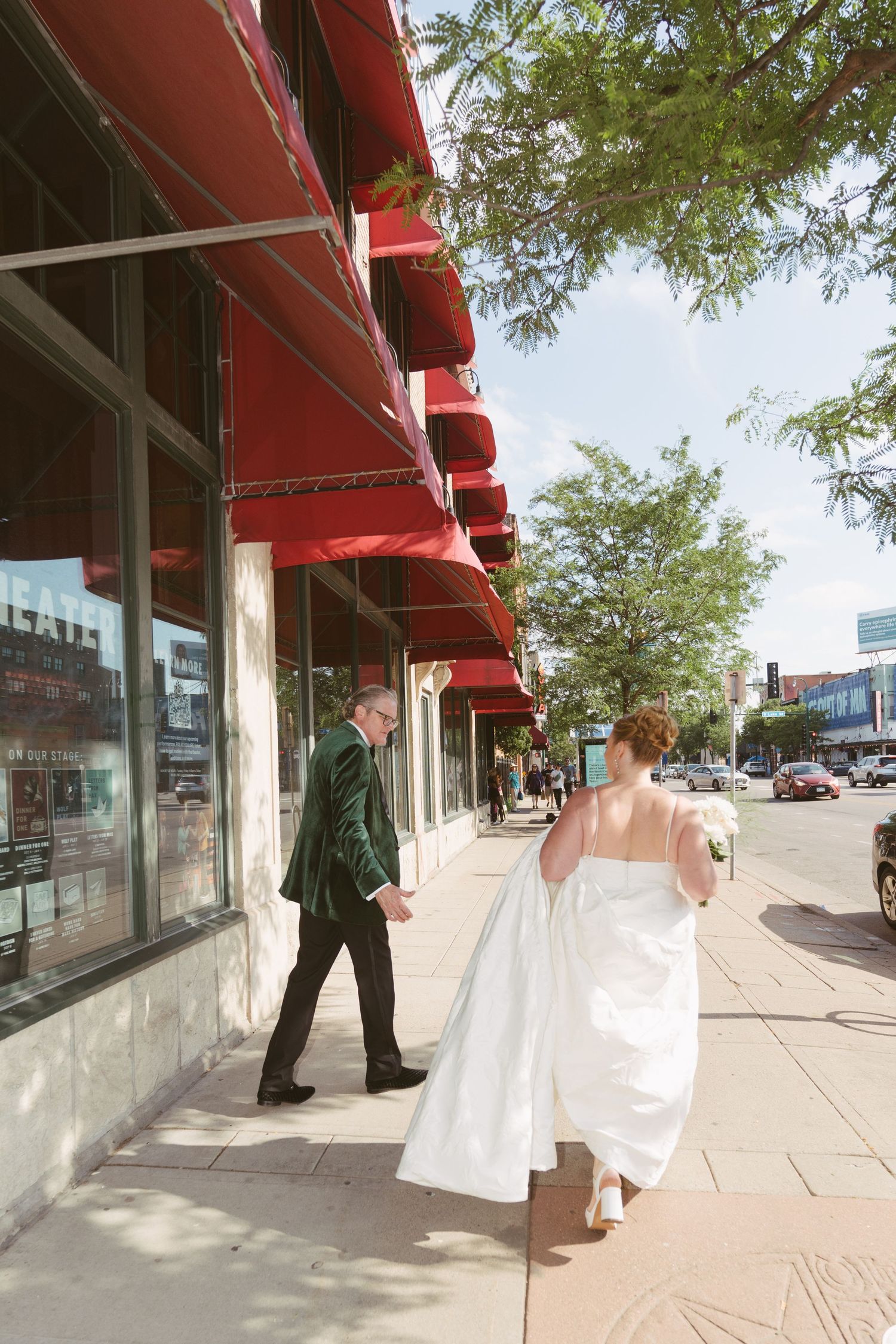 Bride and groom walking hand in hand along a sunny urban sidewalk with red awnings.