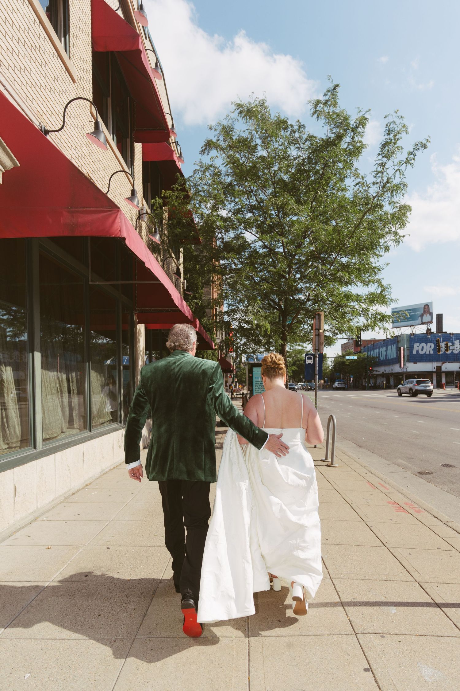 Bride and groom walk hand-in-hand down a sunny urban sidewalk, groom in green velvet jacket with red shoes.