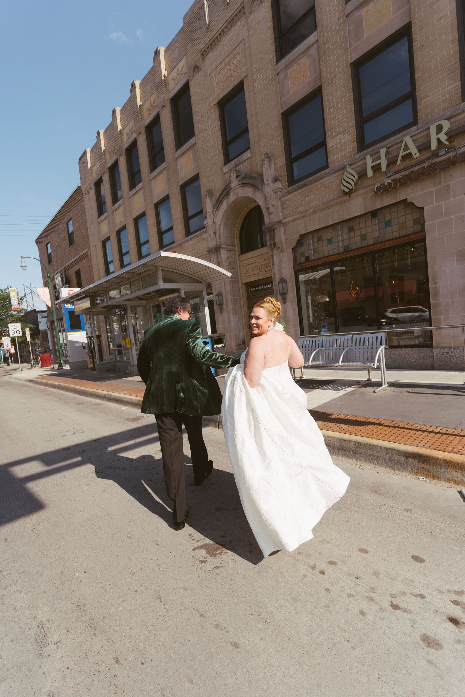 Bride in white gown and groom in dark suit walk together along a sunny urban street past a brick building.