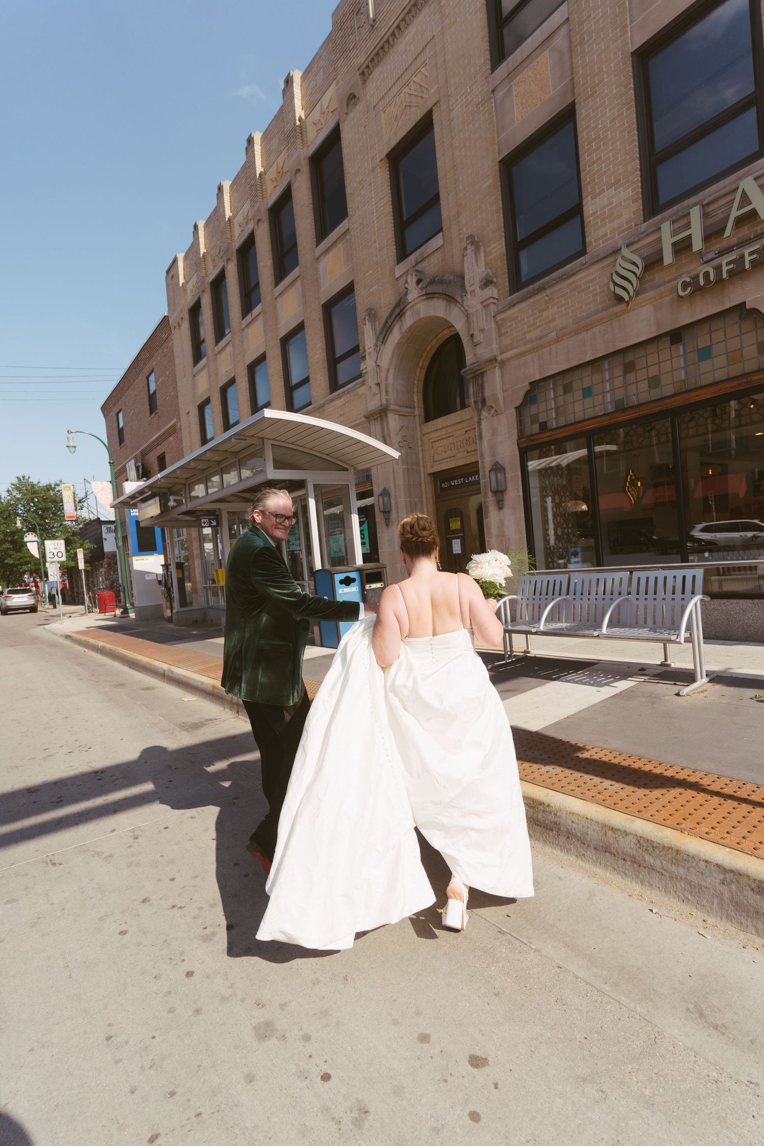 Bride in white gown and groom in green tuxedo walk along sunny urban street.