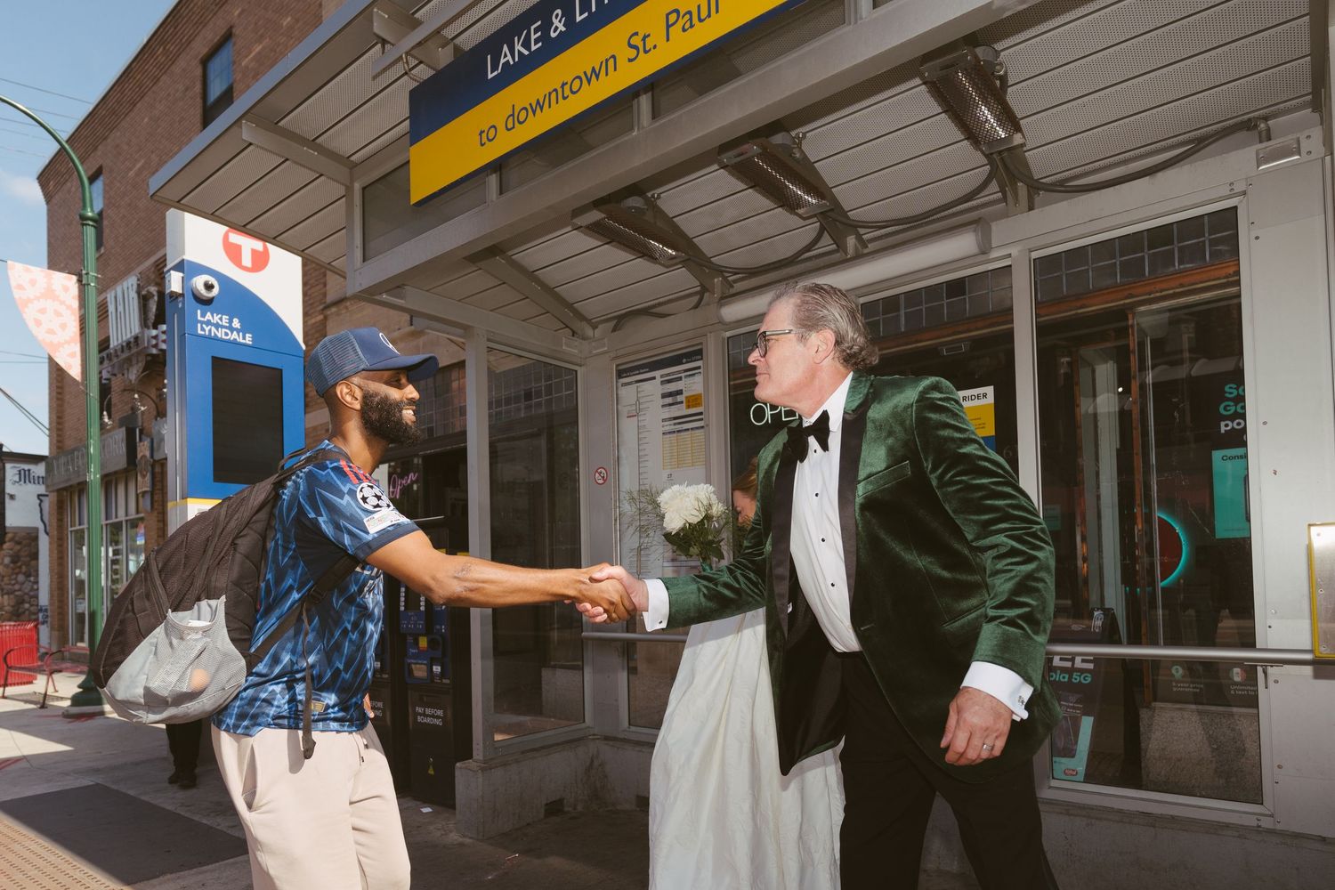 Groom in green velvet tuxedo shakes hands with a stranger outside a Downtown St. Paul Lake & Lyndale transit station.