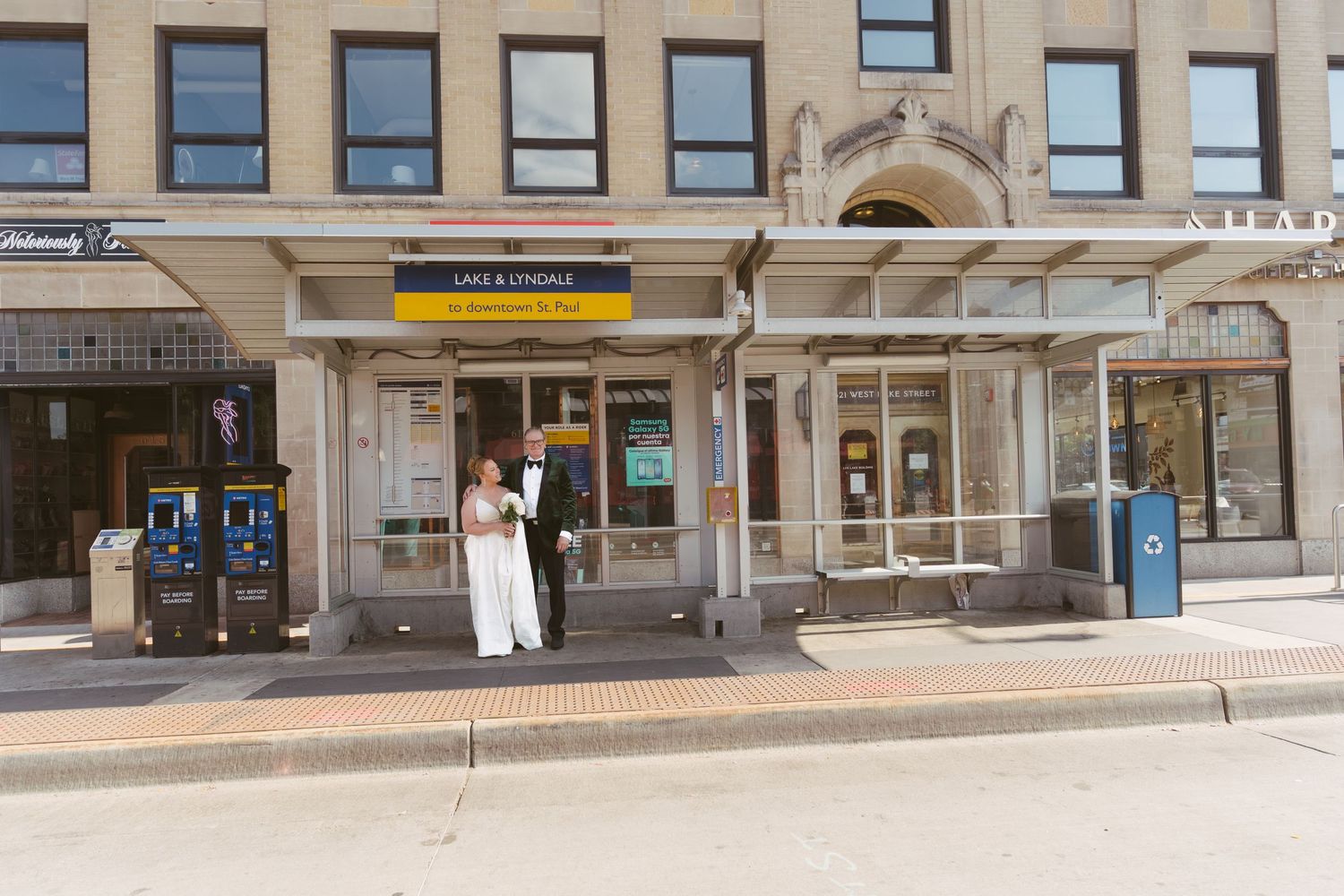 Bride and groom in wedding attire standing outside a Lake and Lyndale transit stop on a city street.