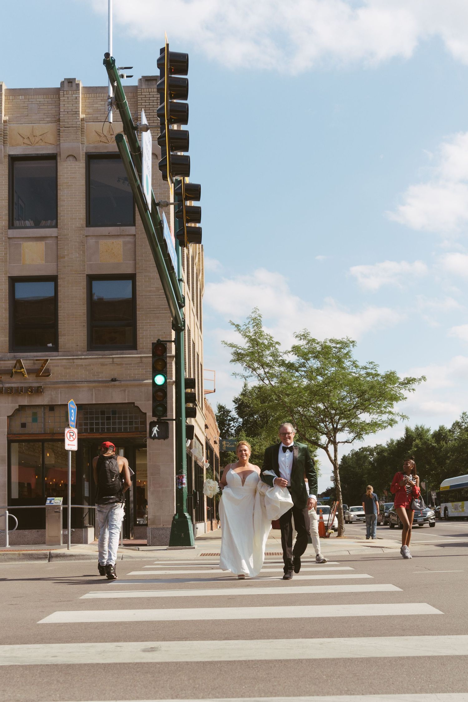 Bride and groom crossing a city street crosswalk in formal wedding attire on a sunny day.