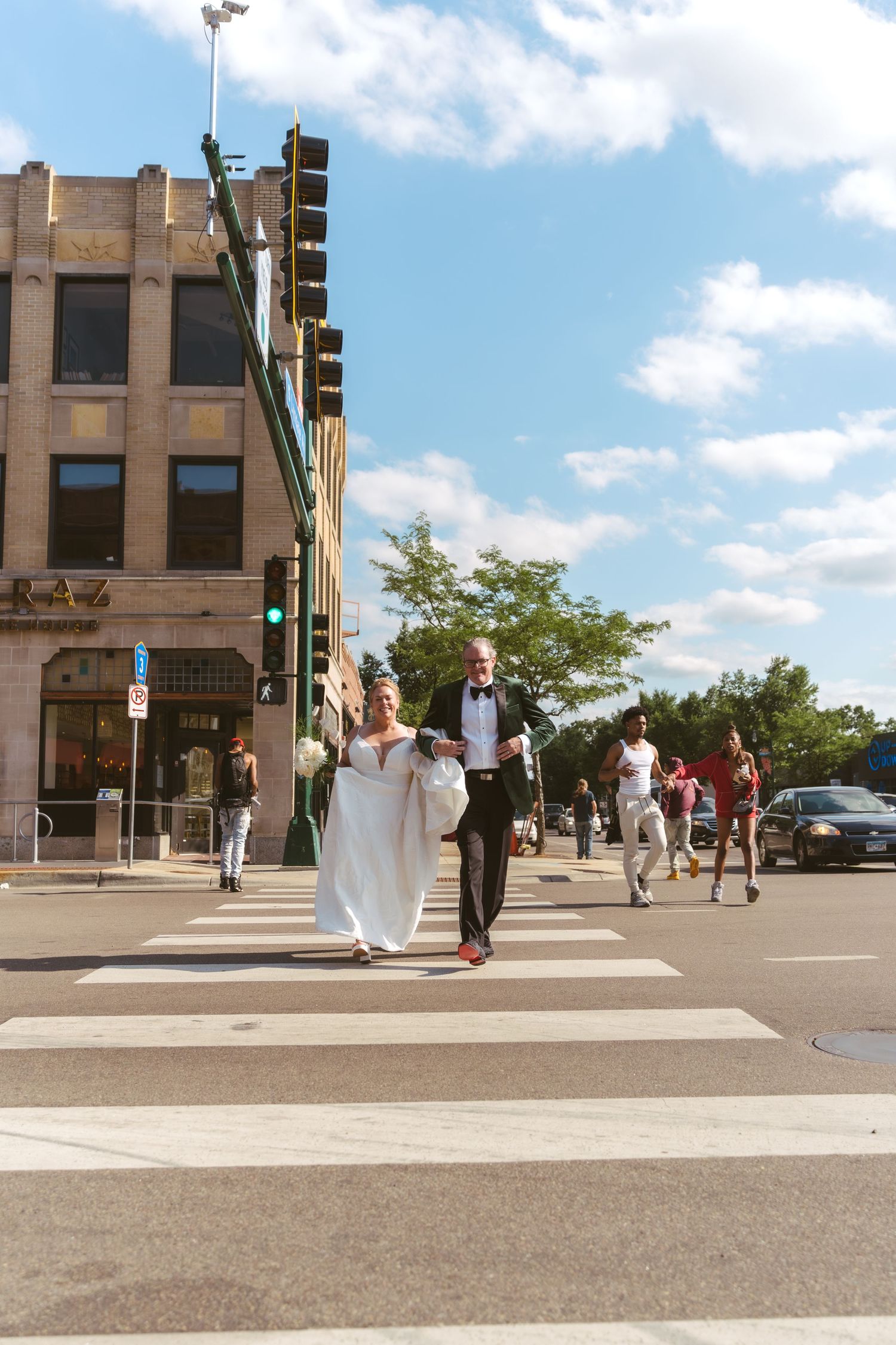 Bride and groom in formal attire cross a busy urban intersection on a sunny wedding day.